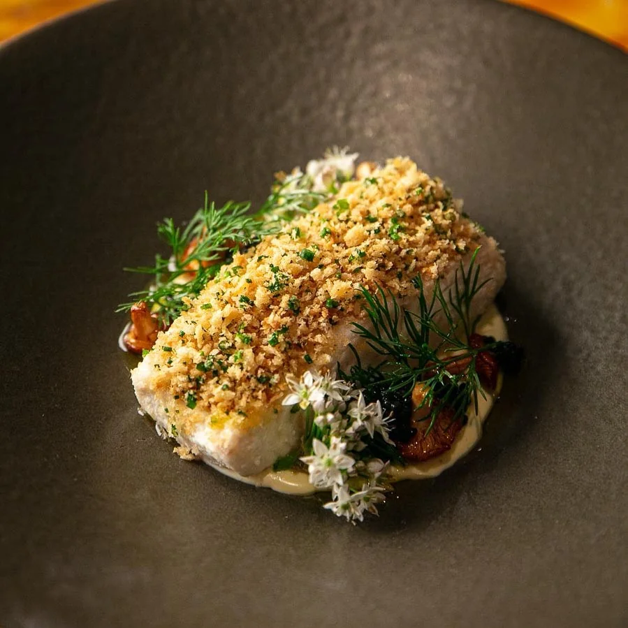 Golden fish with a crumb topping and green herbs on a dark plate