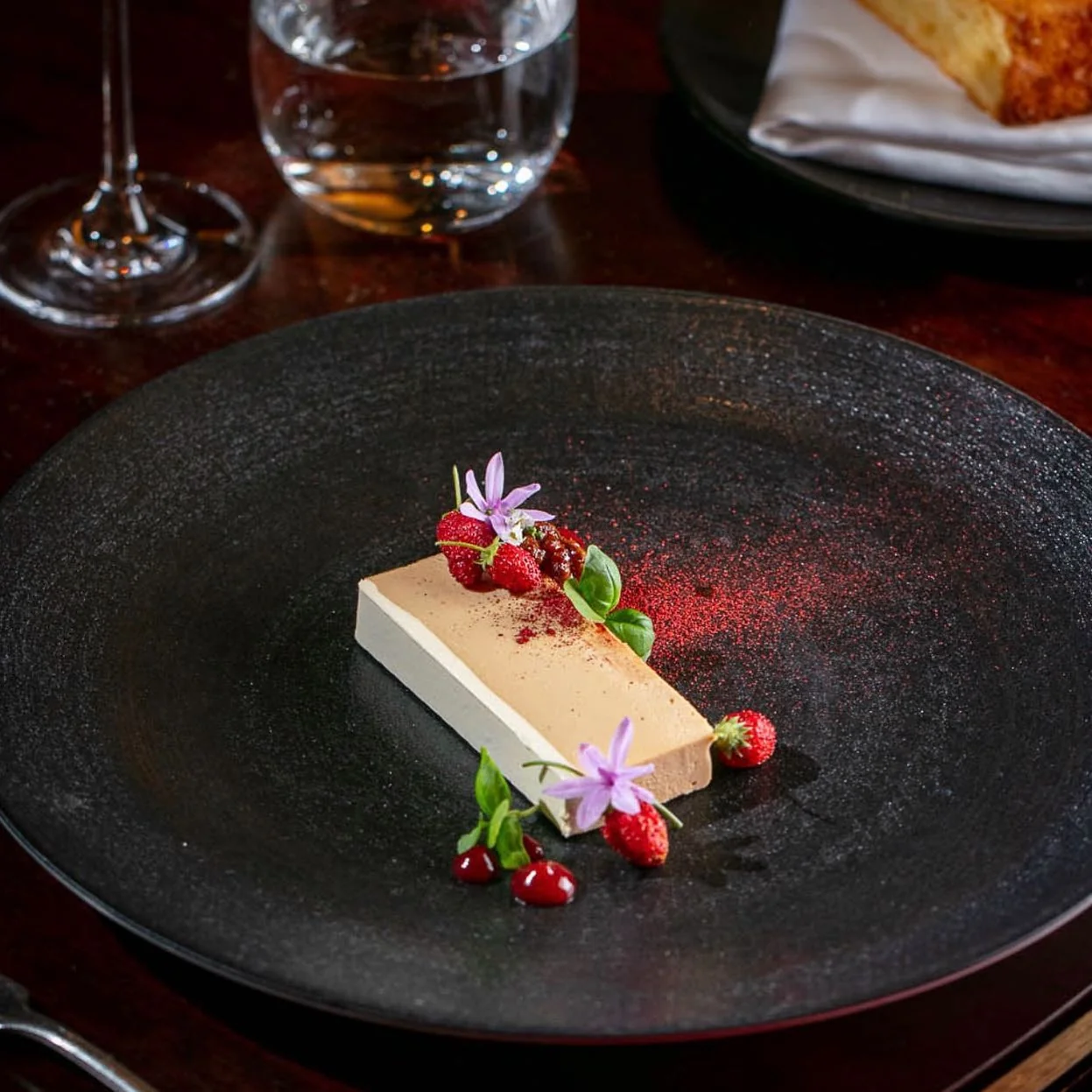 A rectangular parfait with red fruit and flowers on a dark plate